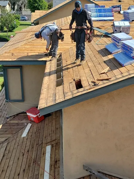 Two workers in protective gear working on the roof of a building, with various construction materials and tools visible.